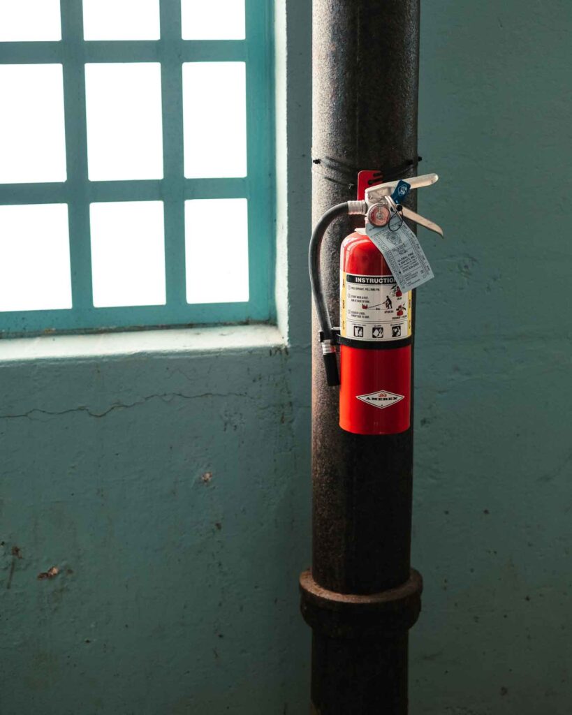 A fire extinguisher connected to a metal pipe in an industrial space, next to a window